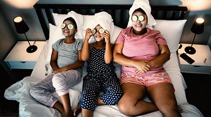 A mom and two young daughters laying on a bed doing fracials with cucumbers on their eyes