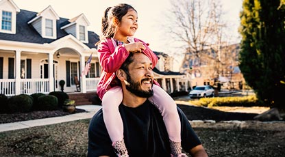 A smiling child sits on their parent's shoulders in front of their new house