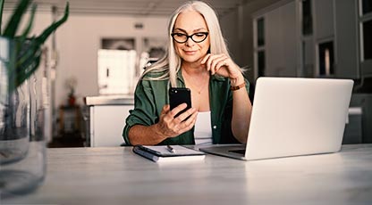 Person wearing glass at desk looking at phone. Computer is on the desk.