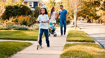 A family walking on a sidewalk with a child riding a scooter and another child asleep in a stroller