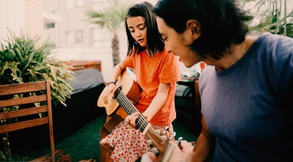 A young person plays the guitar while their older sibling smiles and listens