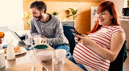 A smiling couple eating breakfast as one looks at a laptop and the other looks at their phone
