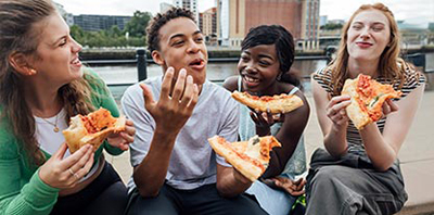 Four friends sit by an urban waterfront, laughing and enjoying large slices of pizza in a relaxed outdoor gathering.