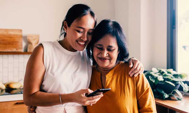 A younger adult has their arm around an older adult as they look at something on their smartphone