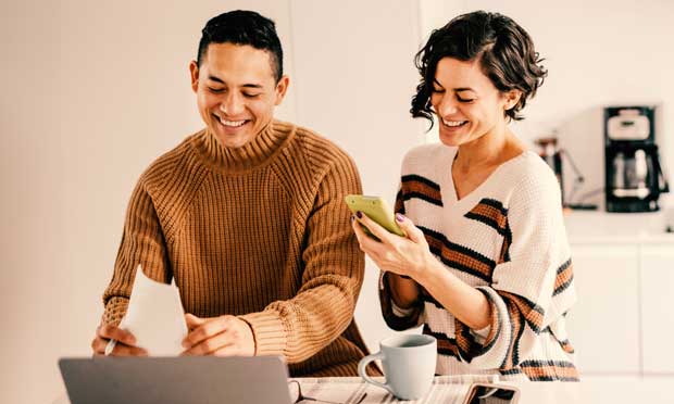 A couple smiling while working on a laptop computer and smartphone