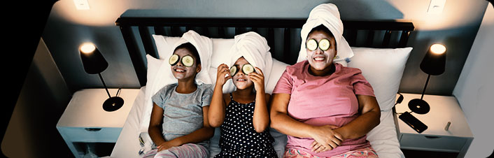 A mom and two young daughters laying on a bed doing facials with cucumbers on their eyes